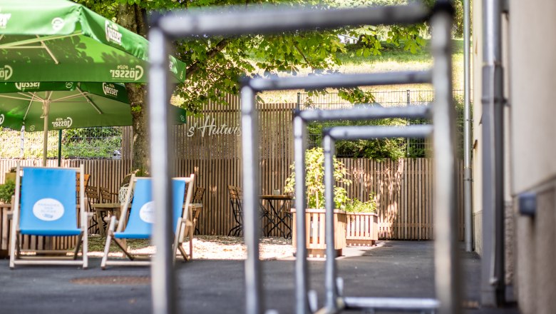 Blick auf einen Gastgarten mit gr&uuml;nen Sonnenschirmen und blauen Liegest&uuml;hlen, umgeben von B&auml;umen und Holzzaun, im Vordergrund Radst&auml;nder.