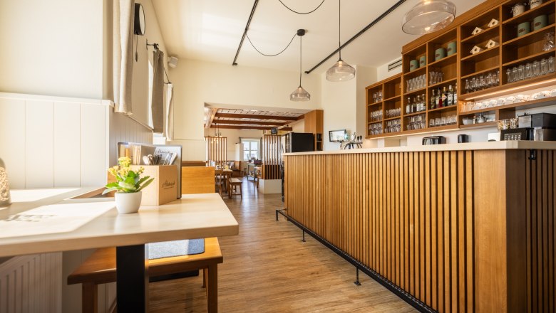 Innenansicht eines modernen Restaurants mit Holzm&ouml;beln und Theke, durch Fenster kommt viel Licht herein, &uuml;ber der Schank h&auml;ngen moderne Lampen.