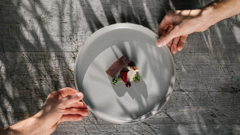 Zwei Hände halten ein Teller mit einem kunstvoll angerichteten Gourmetgericht über einem Holztisch.