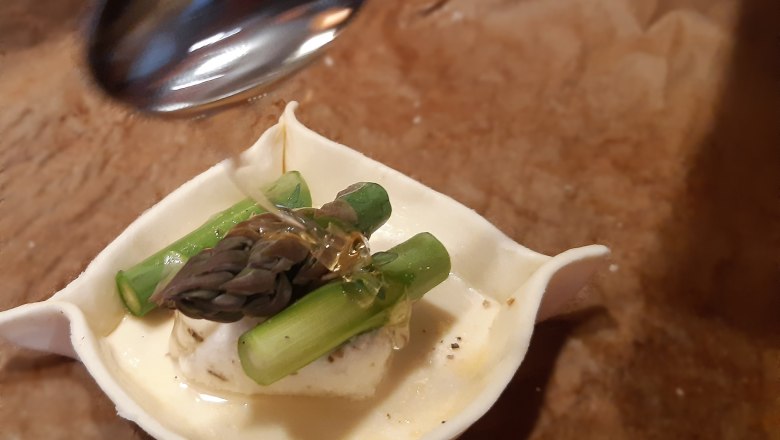 Roher Blättereig mit Ziegenkäse und Spargelstücken, darüber ein Löffel mit Honig.