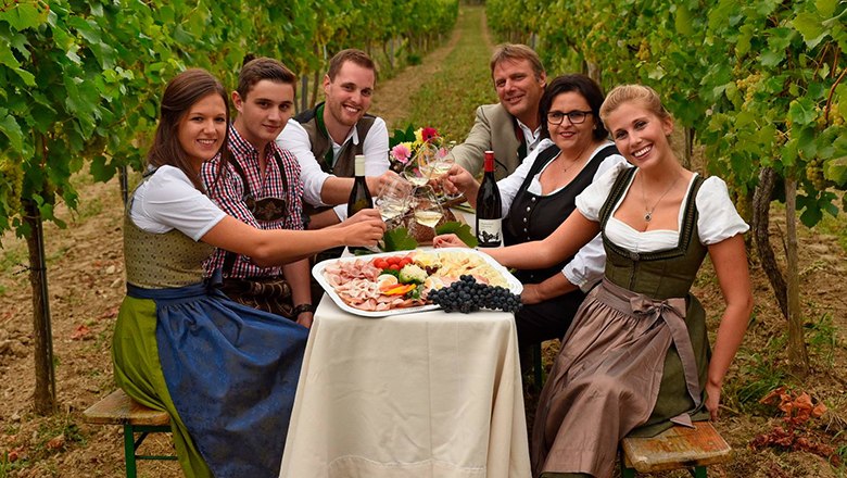 Eine Familie sitzt auf einem Tisch mit Wein, Wurstplatten und Weingläsern zwischen Weinstöcken.