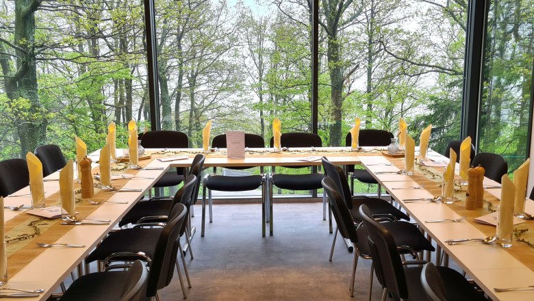 Gedeckte Tische mit gelber Dekoration und schwarze Stühle vor einer großen Fensterfront mit Ausblick auf Bäume.