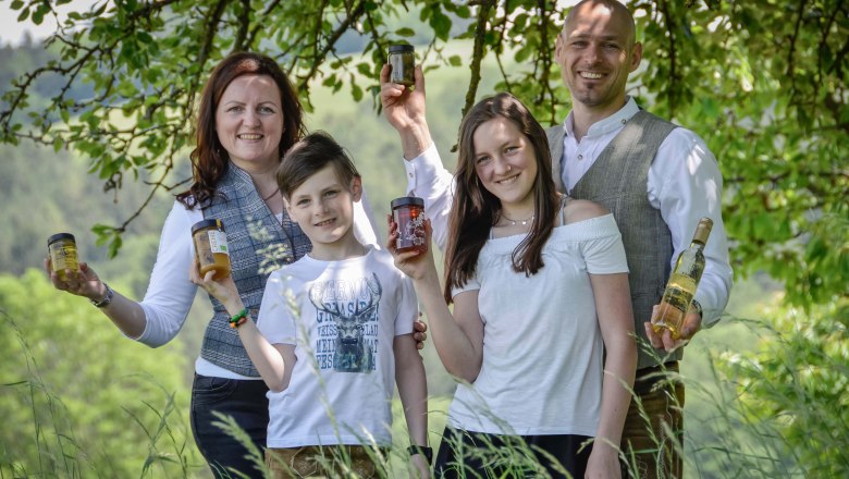 Eine Familie mit zwei Kindern steht auf einer grünen Wiese vor einem Baum und jede Person hält ein Glas Honig in der Hand.