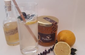 Ein Glas Honiglimonade mit Minze und Zitrone, daneben Honigglas, Zitronen, Kräuter und Sirupflasche.