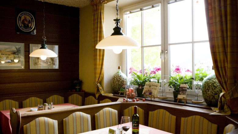 Eine gemütliche Gaststube mit Bänken, Tischen, Lampen und einem großen Fenster mit Vorhängen.