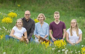 Eine Familie mit drei Kindern sitzt mit zwei Hühnern auf einer grünen Wiese mit gelben Blumen.
