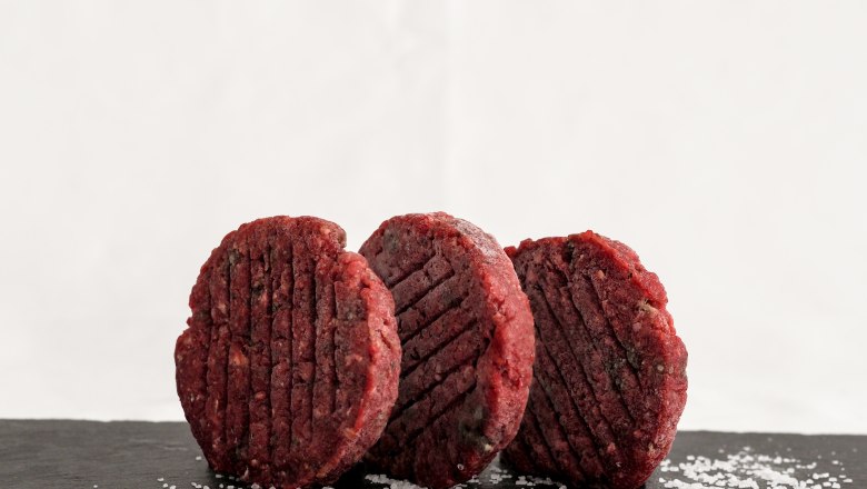 Drei rohe Burgerpatties befinden sich stehend auf einer dunklen Schieferplatte, daneben ist Salz.