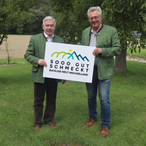 Zwei Männer in Tracht mit Schild ‚Sooo gut schmeckt Bucklige Welt–Wechselland‘.