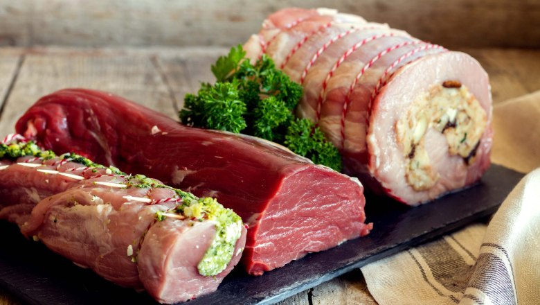 Verschiedene Fleischstücke auf einem Holzbrett, dekoriert mit Kräutern.