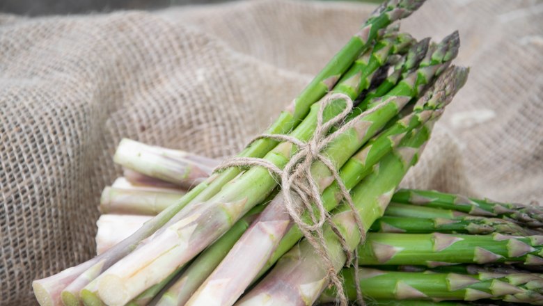 Zwei Bündel grüner Spargel, mit Schnur gebunden, auf braunem Jutestoff liegend.