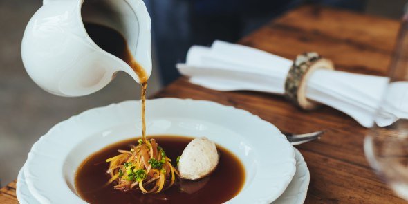 Ein Teller mit Suppe, Gemüse und Knödel, eine Hand gießt mit einer Kanne Suppe in das Teller.