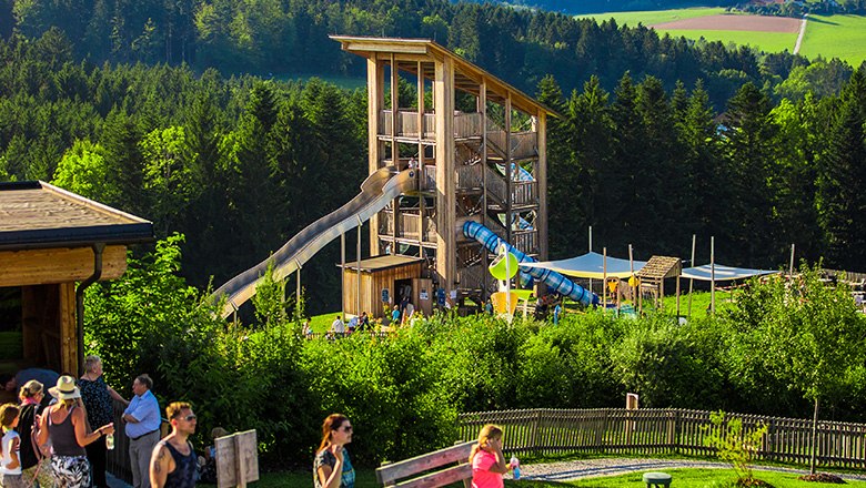 Rutschenturm aus Holz umgeben von Bäumen, und Menschen im Vordergrund.