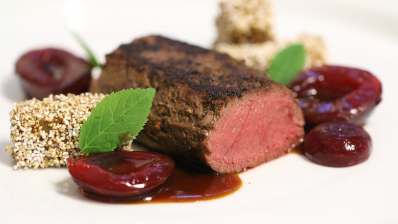 Gourmetgericht mit rosa gebratenem Fleisch, Obst und Kräutern auf einem weißen Teller angerichtet.