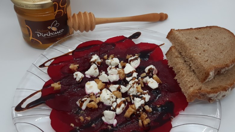 Rote Rüben Carpaccio mit Ziegenfrischkäse, Nüssen, Balsamico und Brot auf Glasteller, daneben Honig.
