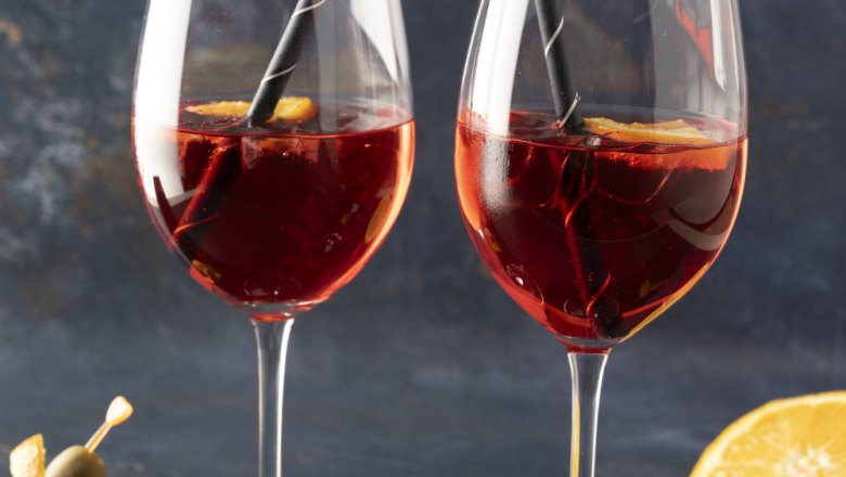 Zwei Weingläser mit Martini Vibrante Sprizz, Orangen und Strohhalmen, daneben Spieße mit Orangen und Oliven.
