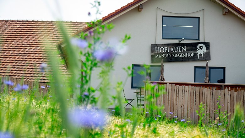 Außenansicht Hofladen Mandl's Ziegenhof mit Holzzaun und Blumen im Vordergrund.