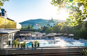 Au&szlig;engel&auml;nde eines Hotels mit Pool, Sonnenschirmen und Liegen, umgeben von Natur und Kirche im Hintergrund.