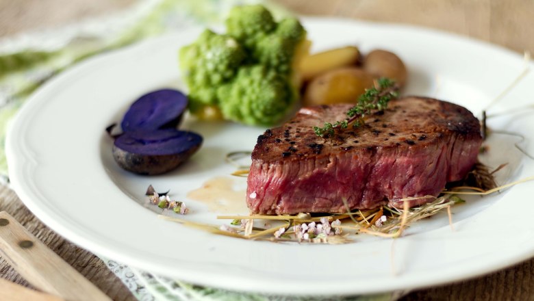 Ein Stück Steak auf einem weißen runden Teller mit Gemüse, Kartoffeln und Kräutern.