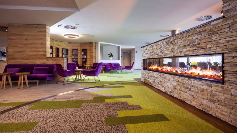 Moderne Hotellobby mit grün-braunem Teppichboden, Kamin, lila Sofas und Stühlen und Holztischen.