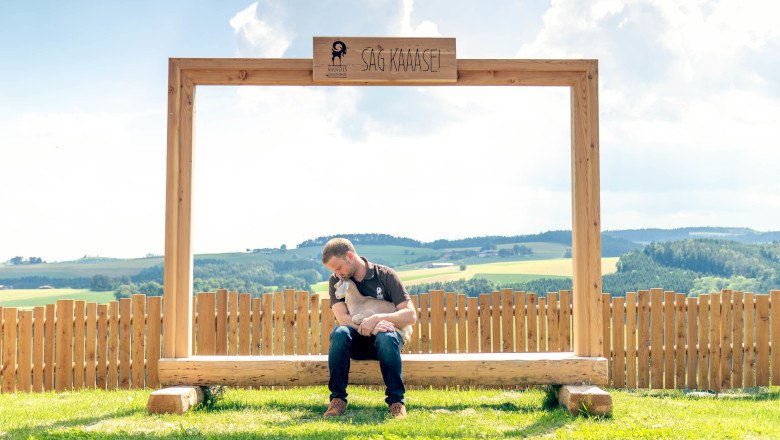 Ein Mann sitzt auf einer Holzbank unter einem großen Rahmen mit der Aufschrift 'SAG KÄÄÄSE' und hält ein Zicklein. Im Hintergrund ist eine ländliche Landschaft zu sehen.