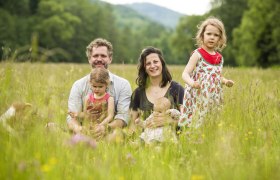 Fr&ouml;hliche Familie mit drei Kindern sitzt auf einer Blumenwiese mit B&auml;umen im Hintergrund.