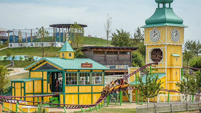 Die Achterbahn "Bucklbahn" im Eis-Greissler Erlebnispark mit gelben Gebäuden und einem grünen Hügel im Hintergrund.