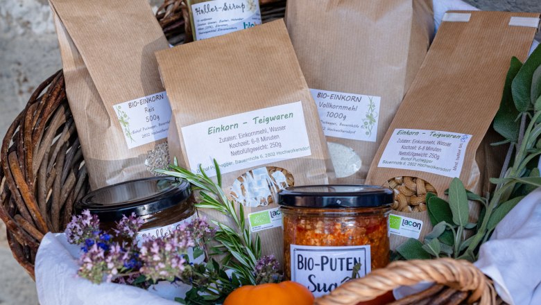 Ein Geschenkkorb mit verschiedenen Produkten wie Einkornwaren, Bio-Puten Sugo und Hollersaft, dekoriert mit Kräutern und Blumen.