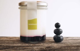 Ein Glas gefüllt mit Joghurt und Fruchtspiegel auf einem Holztisch, daneben 3 gestapelte Heidelbeeren.
