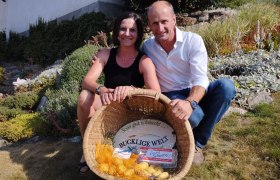 Eine Frau und ein Mann sitzen l&auml;chelnd in einem Garten und halten einen Korb mit Kartoffeln und Eiern.