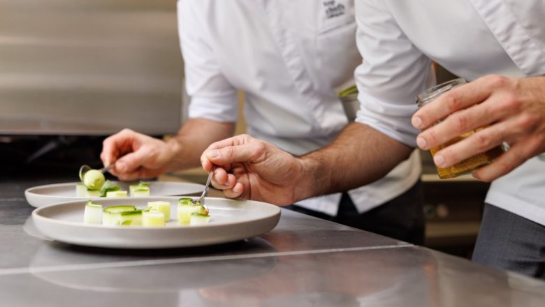 Zwei Köche in weißen Kochjacken richten Gourmetgerichte auf weißen Tellern an.