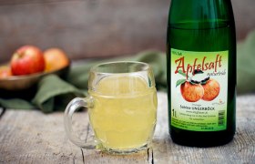 Eine Flasche Apfelsaft und ein Glas gefüllt mit Apfelsaft auf einem Holztisch, im Hintergrund ein grünes Tuch.
