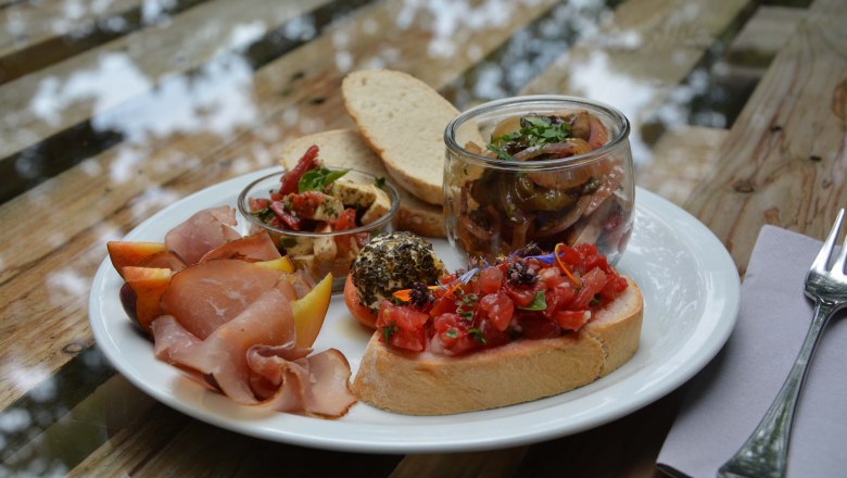 Ein Teller mit Brot, Schinken, Antipasti, Bruschetta und Obst auf einem Holztisch.
